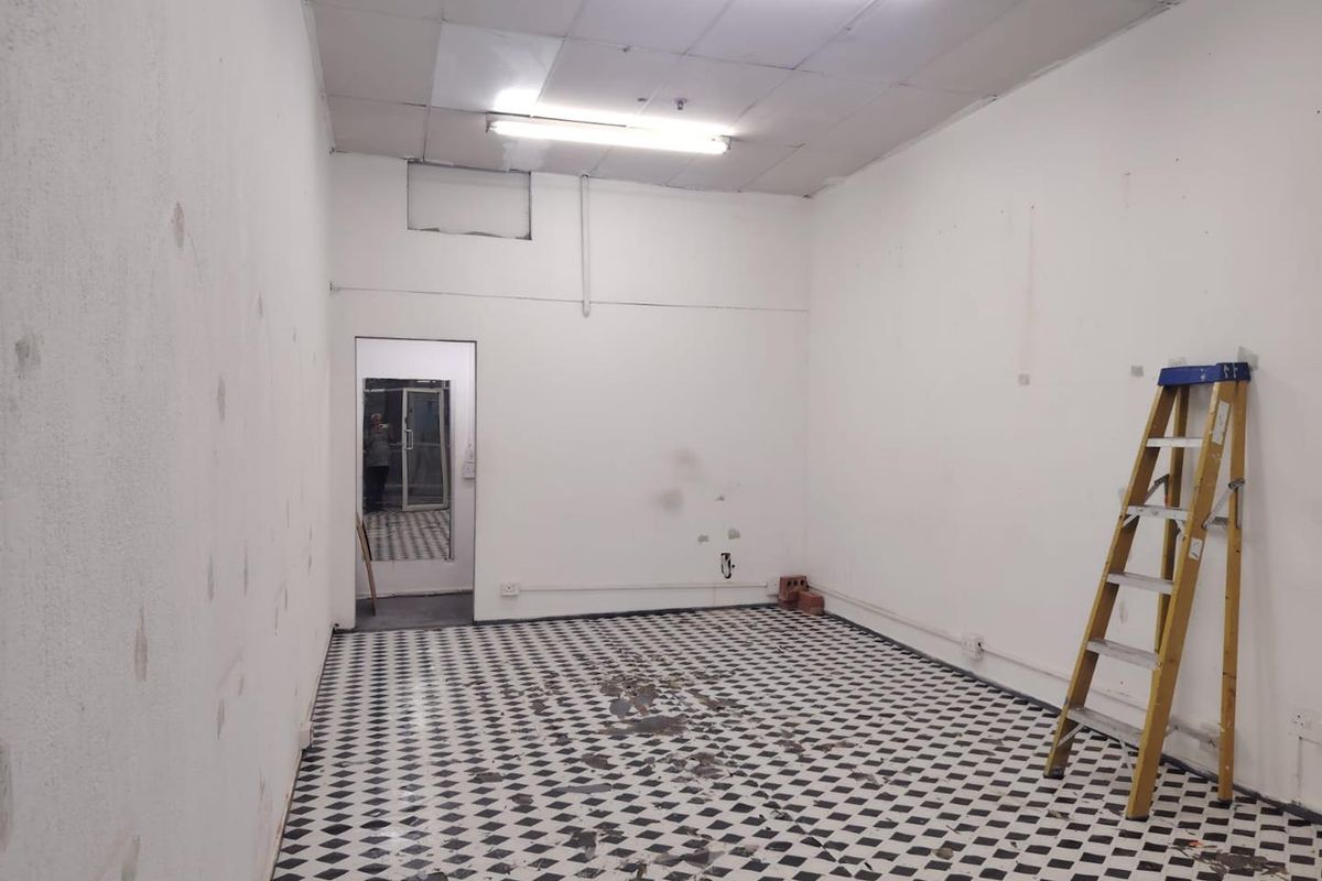 An empty retail space featuring checkered floor tiles, suspended ceiling with fluorescent lighting, and plain white walls requiring cosmetic attention.