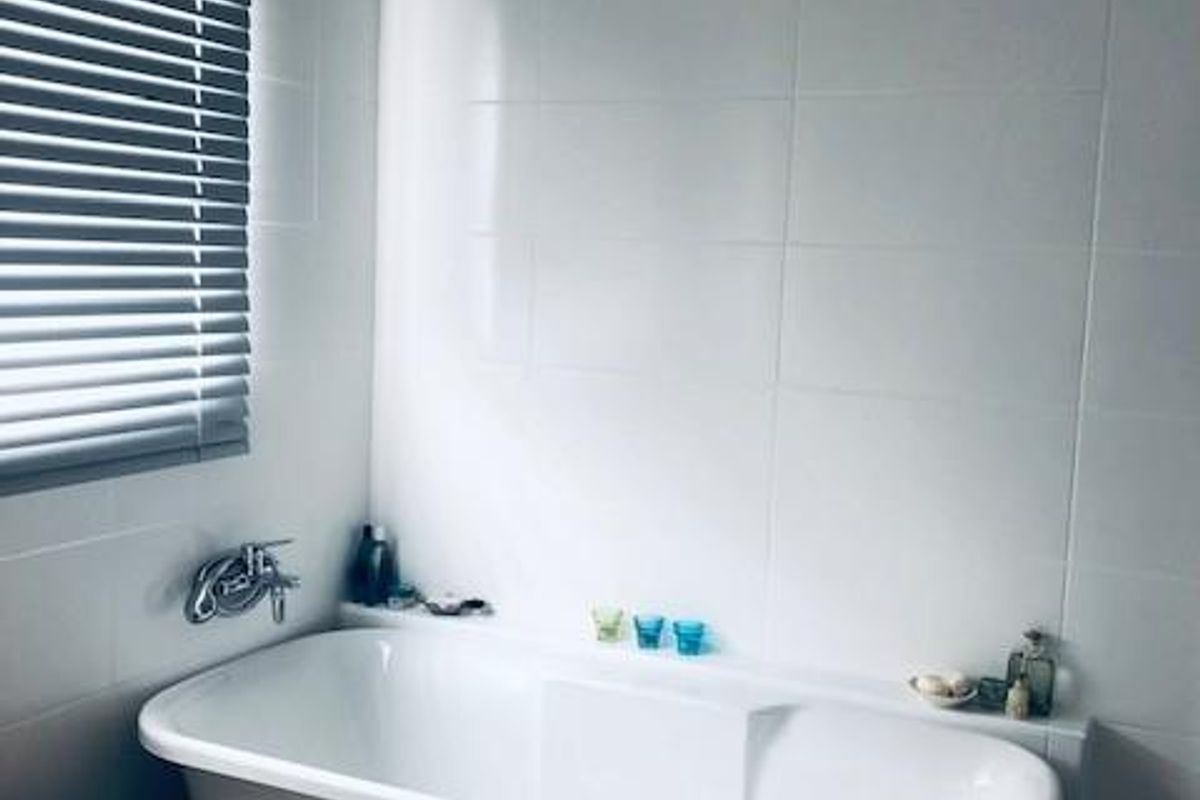A clean, modern bathroom featuring a white freestanding bathtub, wall-mounted chrome faucet, and neutral tiled walls with a window blind.