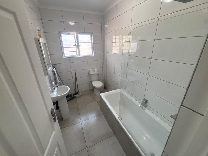 A clean, functional bathroom featuring a pedestal basin, toilet, and a bathtub with a glass shower screen, finished with neutral wall and floor tiles.