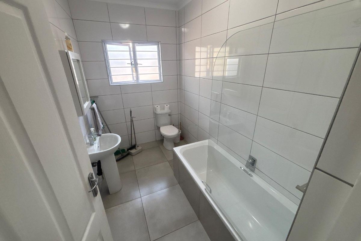 A clean, functional bathroom featuring a pedestal basin, toilet, and a bathtub with a glass shower screen, finished with neutral wall and floor tiles.