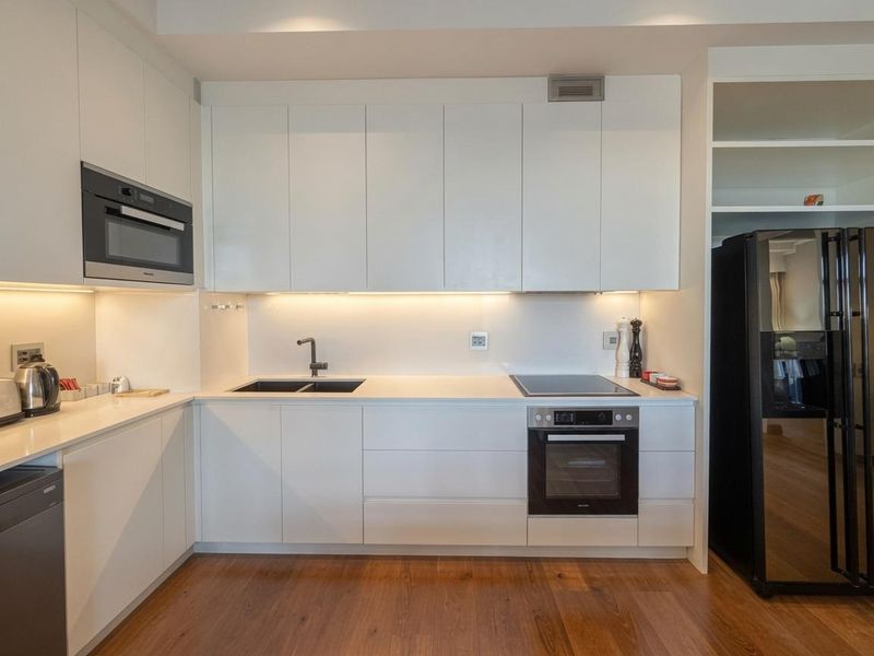 Modern kitchen featuring sleek white cabinetry, integrated appliances, stone countertops, and warm hardwood flooring with efficient under-cabinet lighting.