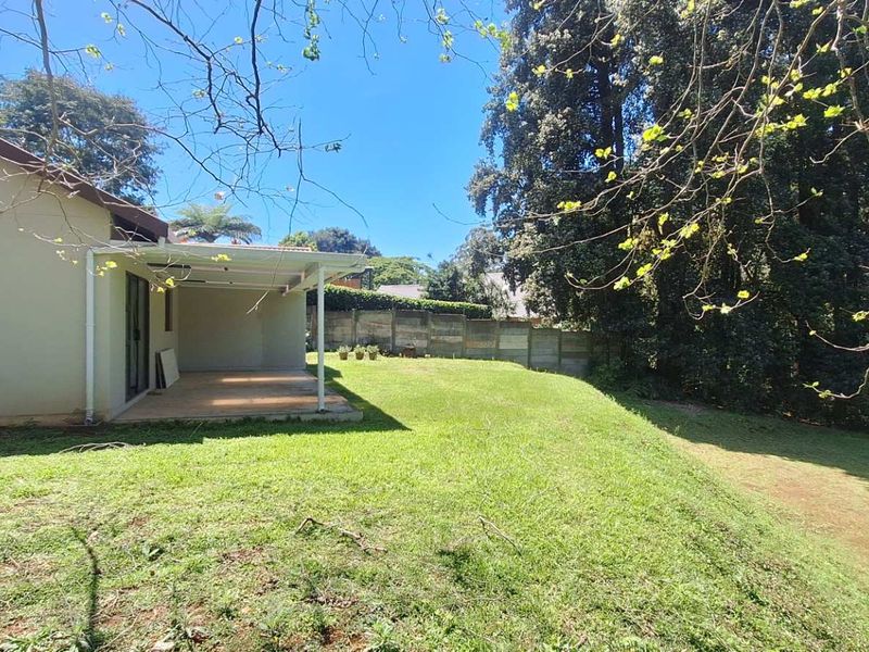 A spacious rear garden featuring a covered patio area and a sloped lawn, bordered by a concrete boundary wall.