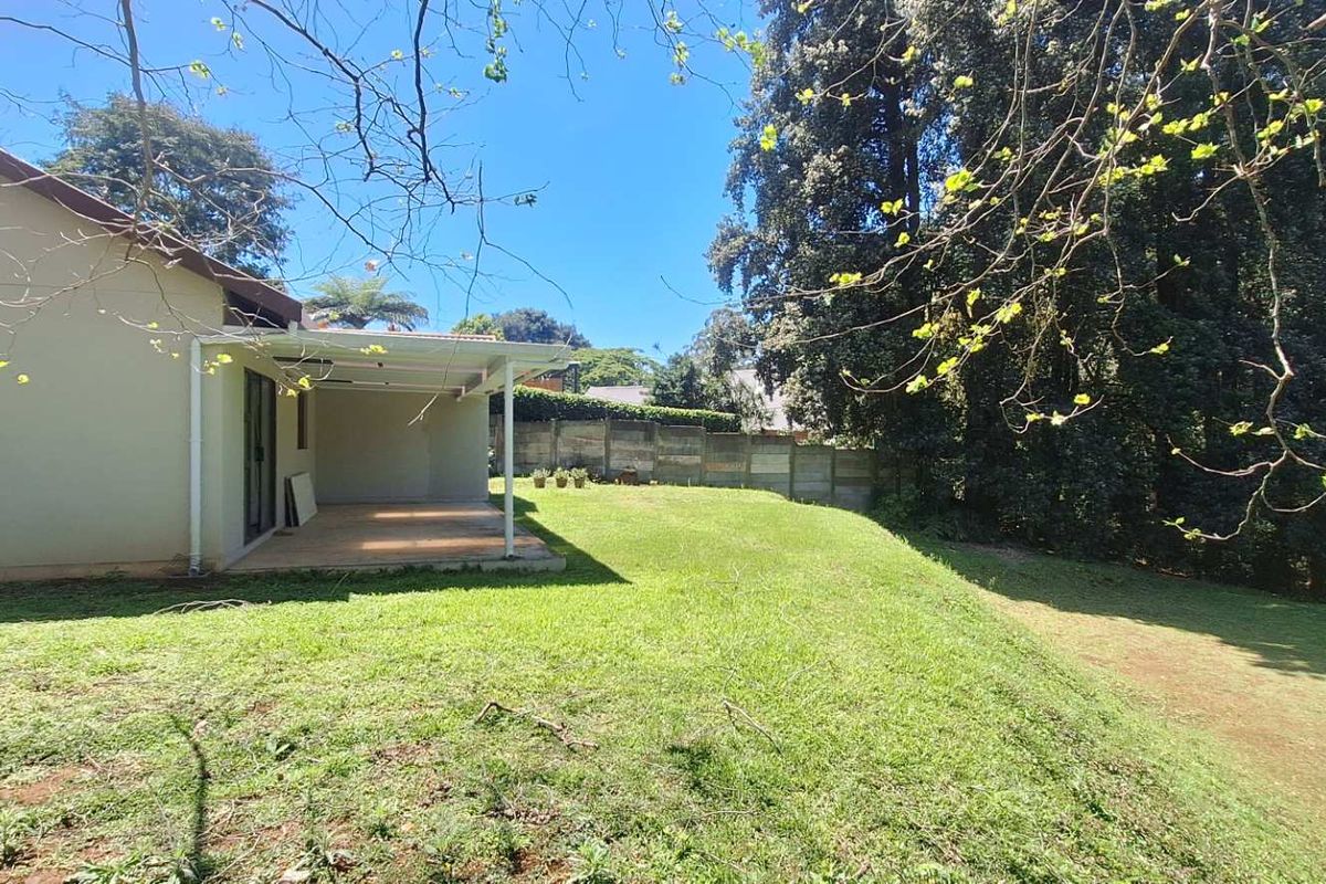 A spacious rear garden featuring a covered patio area and a sloped lawn, bordered by a concrete boundary wall.
