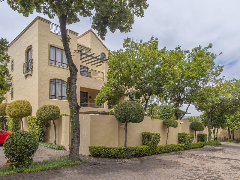 Exterior view of a multi-story apartment building featuring a stucco facade, balconies with wrought iron railings, and a paved driveway with landscaped garden borders.