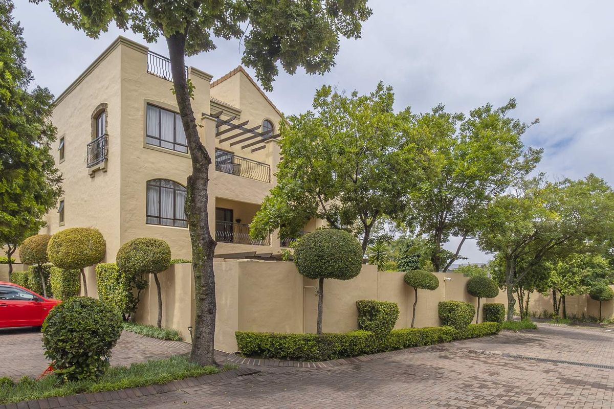 Exterior view of a multi-story apartment building featuring a stucco facade, balconies with wrought iron railings, and a paved driveway with landscaped garden borders.