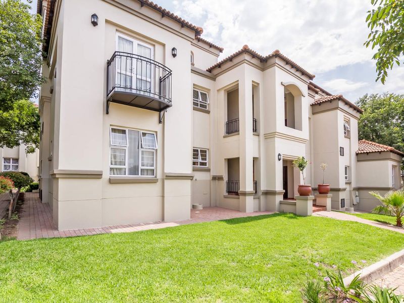 A well-maintained apartment building exterior featuring a landscaped lawn, paved pathways, and classic architectural details including wrought iron balconies and tiled roofing.