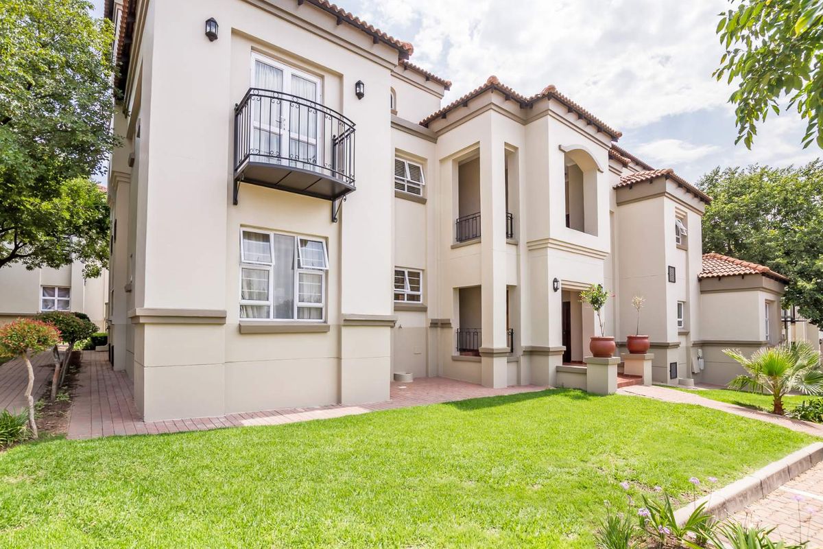 A well-maintained apartment building exterior featuring a landscaped lawn, paved pathways, and classic architectural details including wrought iron balconies and tiled roofing.