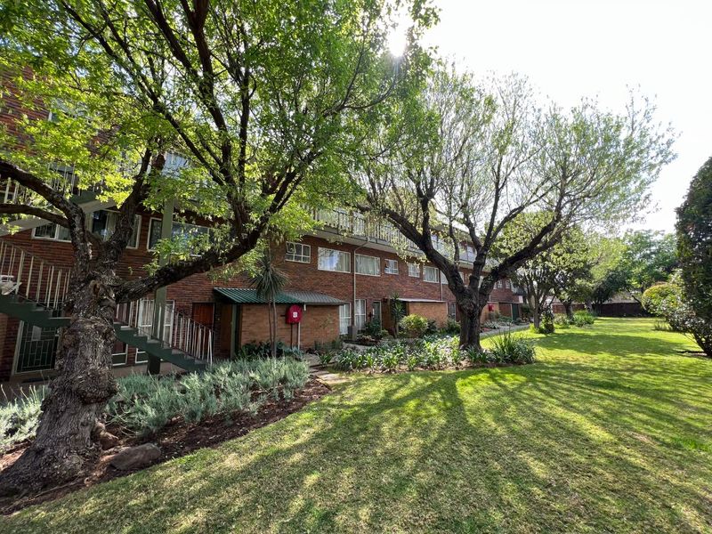 A view of the apartment building's rear facade featuring brick construction, external metal staircases, balconies, and a well-maintained communal garden area.