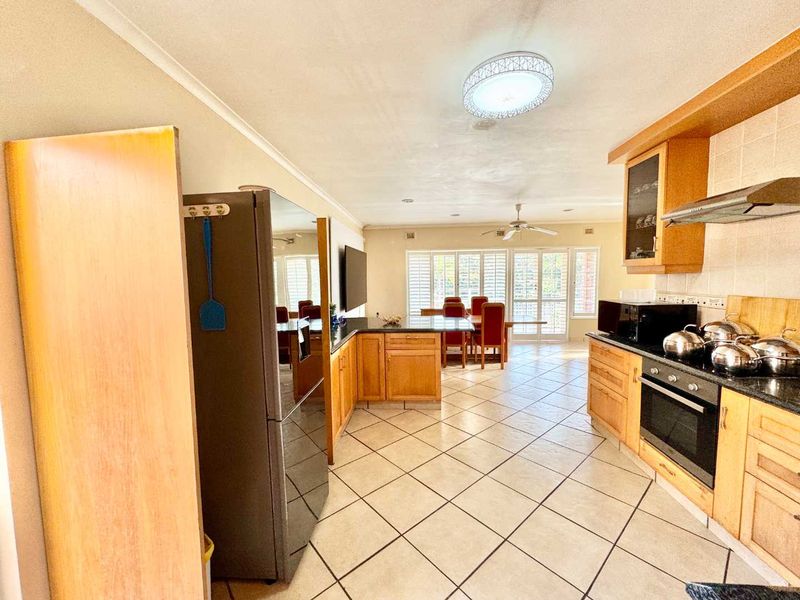 Open-plan kitchen and dining area featuring wooden cabinetry, granite countertops, built-in oven, and tiled flooring with access to the outdoor area via plantation shutters.