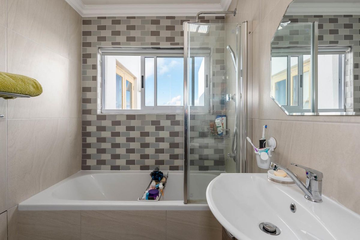 A modern bathroom featuring a combined bathtub and shower unit, tiled walls with a decorative mosaic accent, and a wall-mounted basin.