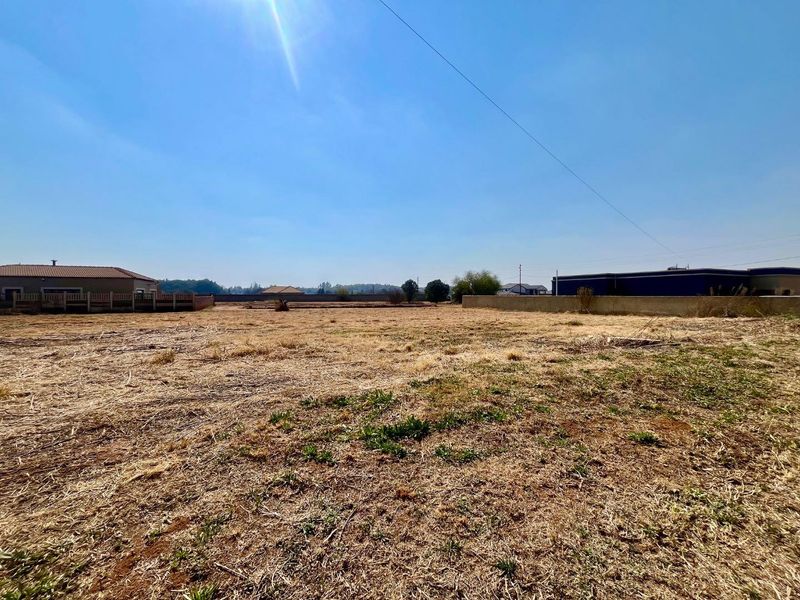 A wide-angle view of a vacant residential plot featuring dry grass, perimeter boundary walls, and surrounding neighborhood structures.