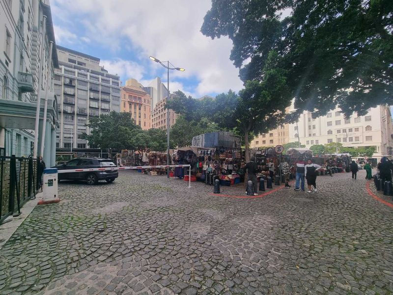 A street-level view of a cobblestone urban square featuring surrounding apartment buildings, mature trees, and a security access point.