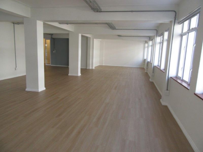 Spacious open-plan office or commercial area featuring light wood-look laminate flooring, structural columns, and large windows providing ample natural light.