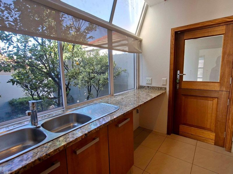A functional scullery area featuring granite countertops, a stainless steel double sink, wooden cabinetry, and direct access to the garden through a timber door.
