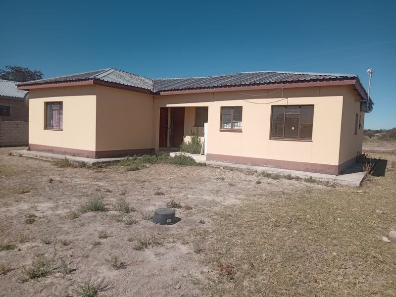 A single-story detached house featuring a corrugated metal roof and simple exterior facade, situated on a large, unlandscaped plot of land.