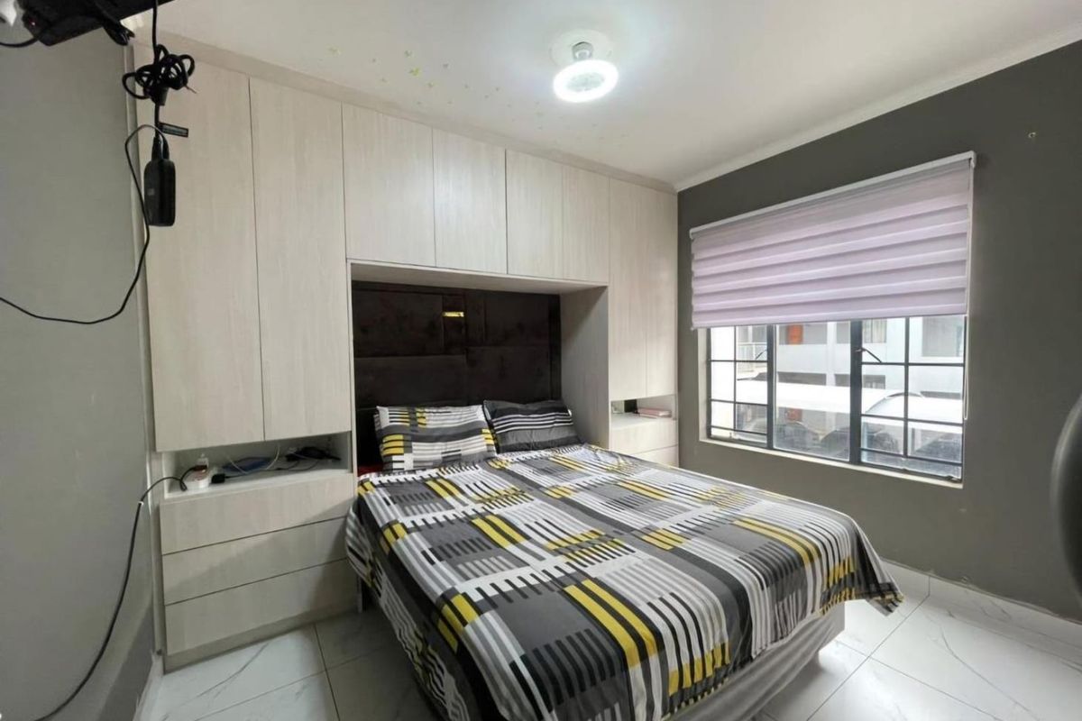 A well-maintained bedroom featuring built-in floor-to-ceiling cabinetry with integrated headboard space, tiled flooring, and a large window with modern roller blinds.