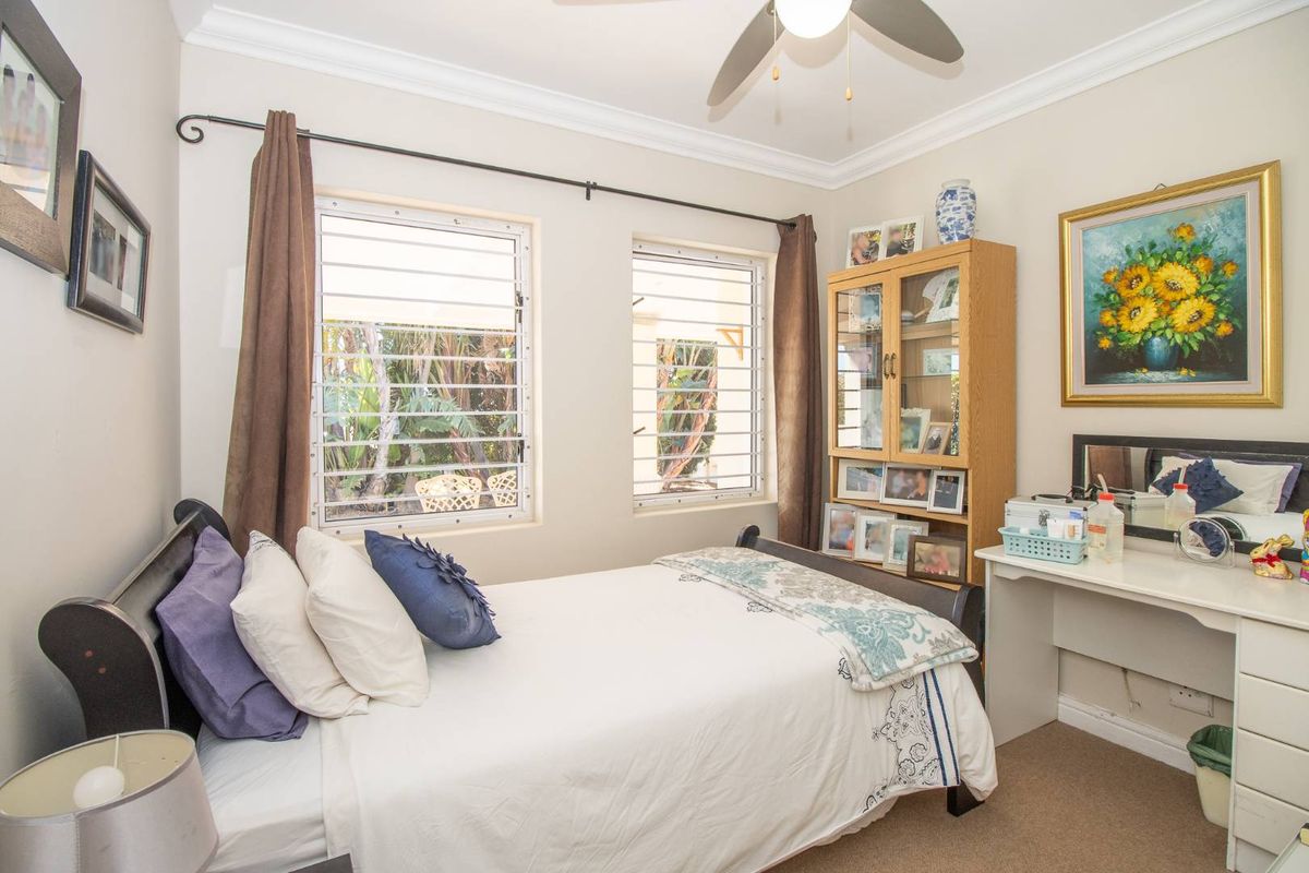 A well-lit bedroom featuring neutral walls, carpeted flooring, ceiling fan, and security-barred windows overlooking the garden.