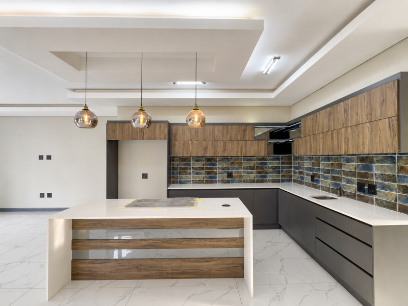 Modern, spacious kitchen featuring a large island, sleek wood-grain and charcoal cabinetry, stone countertops, and a decorative tiled backsplash.
