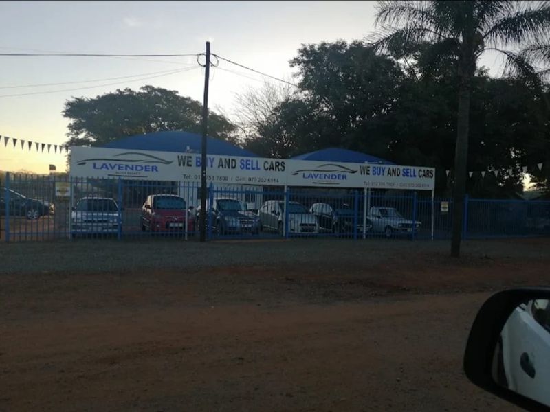 Front view of a car dealership property featuring steel perimeter fencing and covered carport areas for vehicle display.
