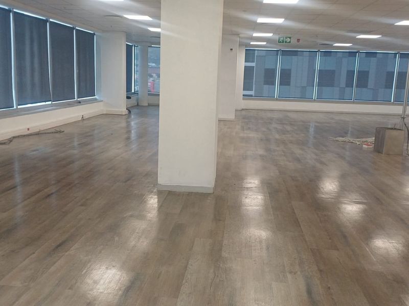 A spacious, open-plan office area featuring modern laminate flooring, large windows with roller blinds, and a clean, neutral aesthetic.