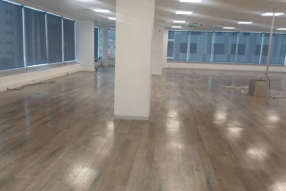 A spacious, open-plan office area featuring modern laminate flooring, large windows with roller blinds, and a clean, neutral aesthetic.