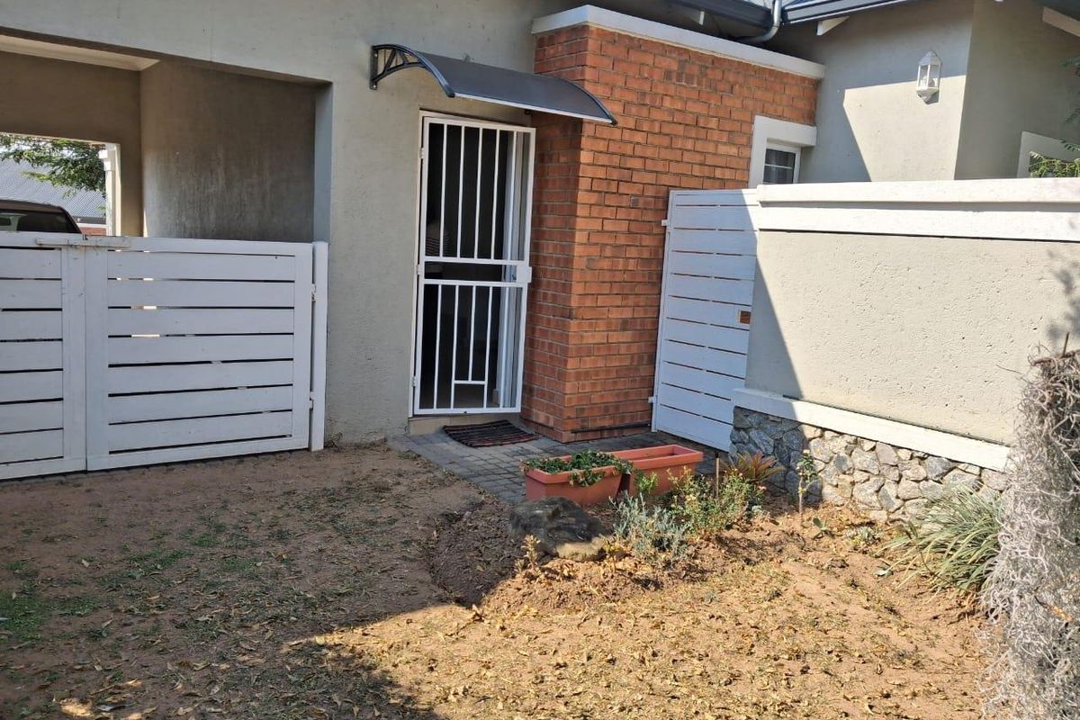 A rear exterior view showing a paved entrance with a security gate, brick detailing, and a small garden area with boundary walling.