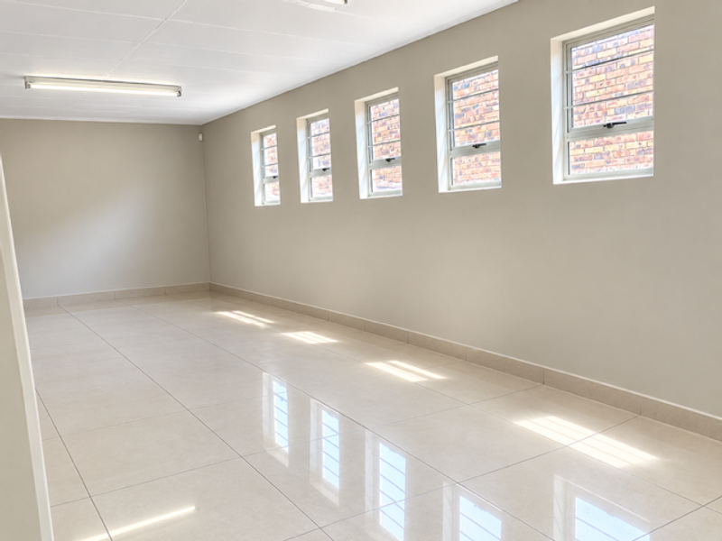 A bright, spacious, and empty industrial office area featuring polished tiled flooring, neutral walls, and multiple steel-framed windows providing natural light.