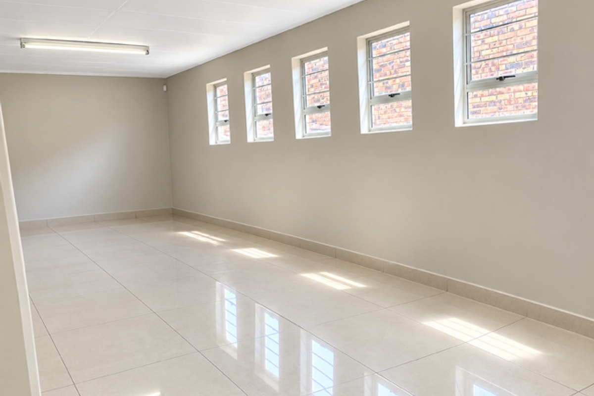 A bright, spacious, and empty industrial office area featuring polished tiled flooring, neutral walls, and multiple steel-framed windows providing natural light.