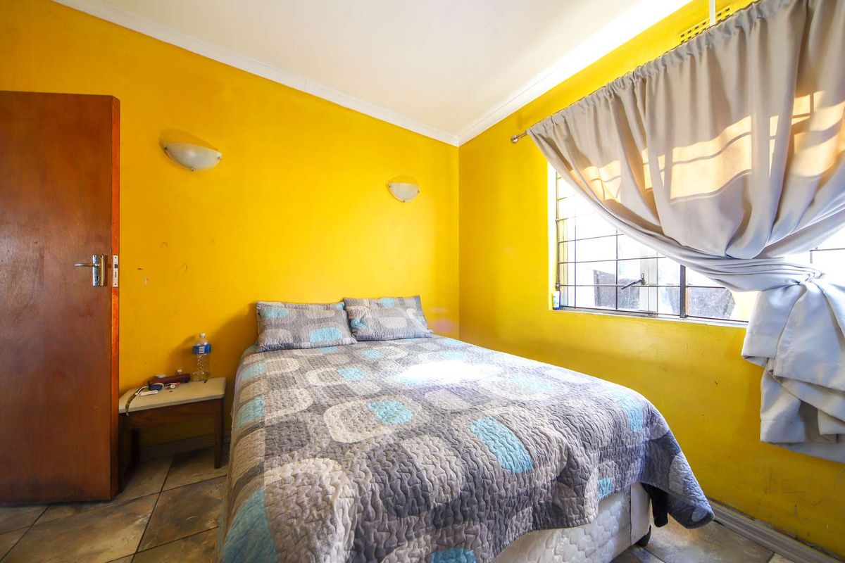 A bedroom featuring bright yellow walls, tiled flooring, and a window with security bars, providing a functional sleeping space.