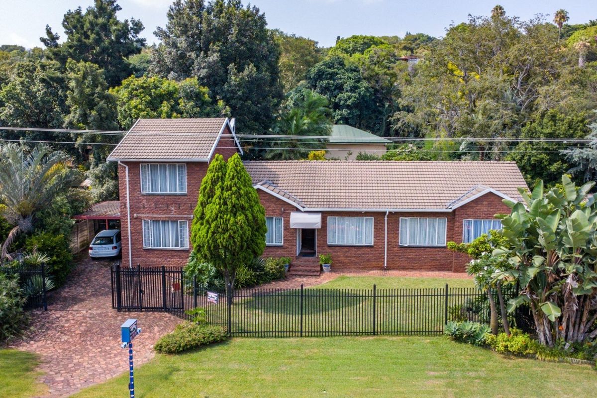 Aerial view of a double-story brick house featuring a tiled roof, paved driveway, and secure perimeter fencing set within a lush, established garden.