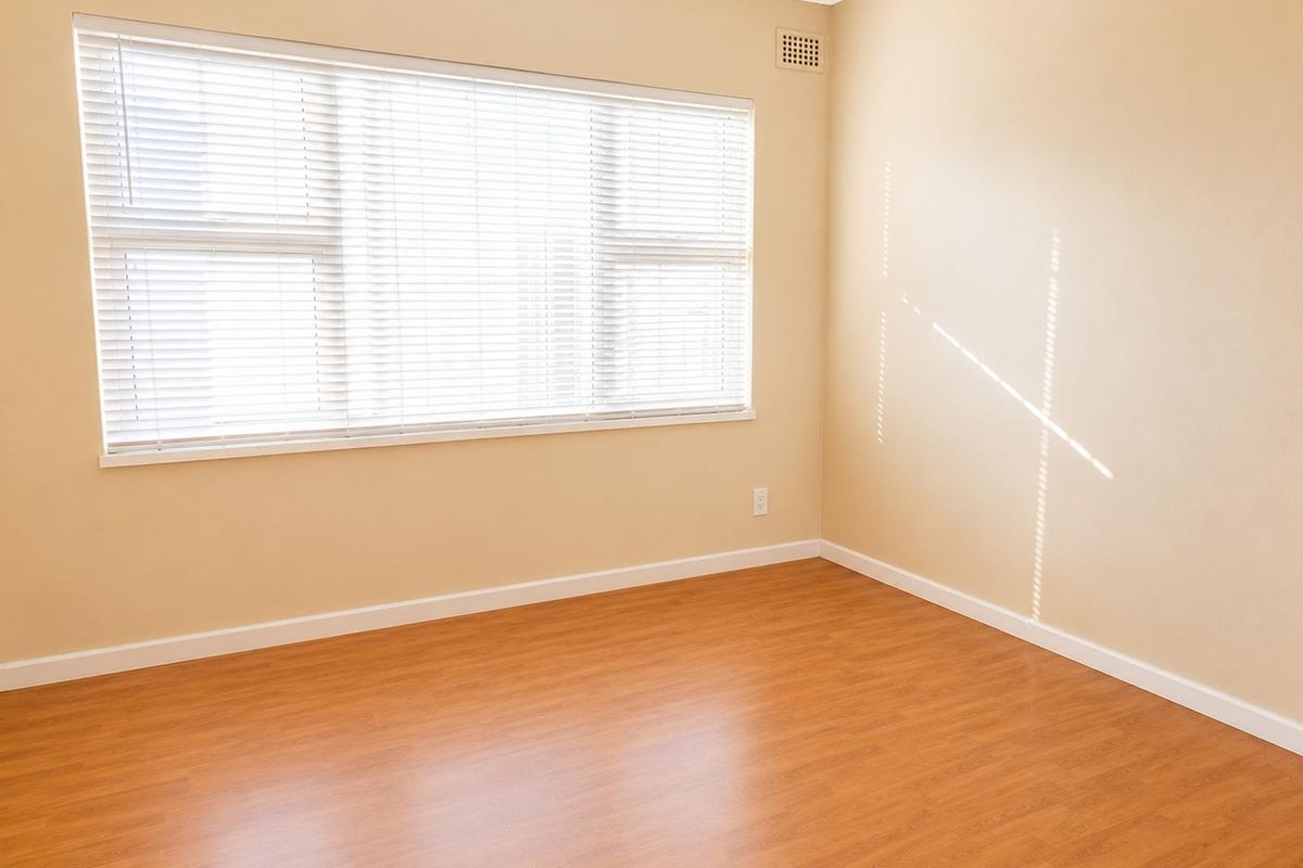 A bright, empty bedroom featuring laminate flooring, neutral walls, and large windows fitted with venetian blinds.