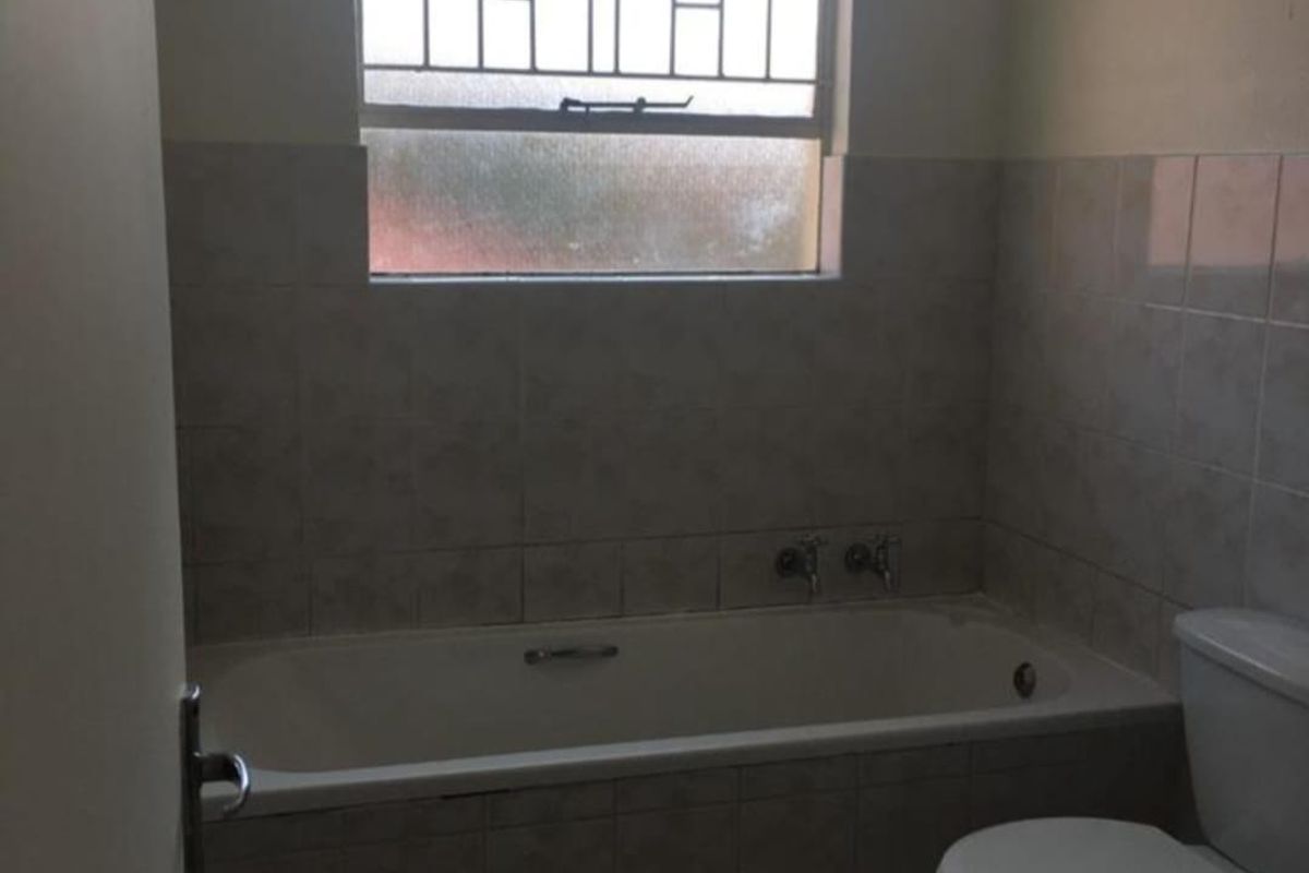 A functional bathroom featuring a built-in bathtub, toilet, and tiled finishes, complete with a window fitted with security bars.