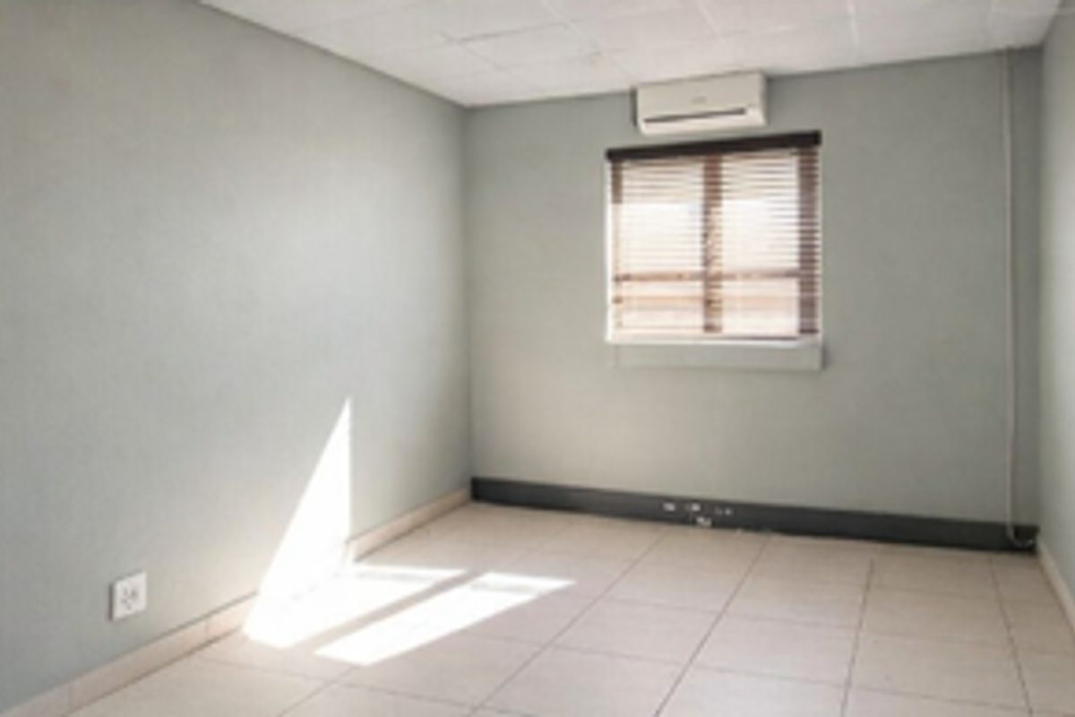 A vacant office space featuring tiled flooring, a drop ceiling, an integrated air conditioning unit, and a window with fitted venetian blinds.