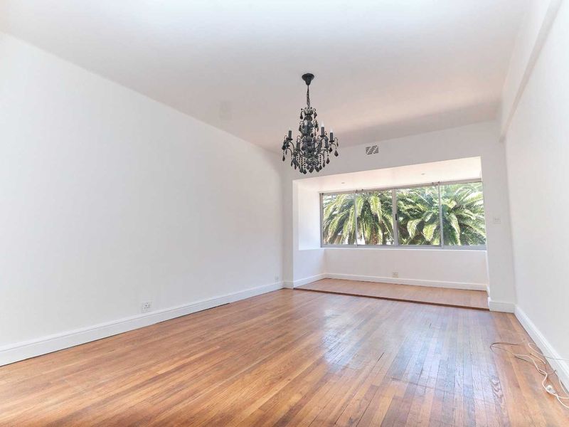 A bright, empty living room featuring polished hardwood flooring, a decorative chandelier, and a large window with a view of palm trees.