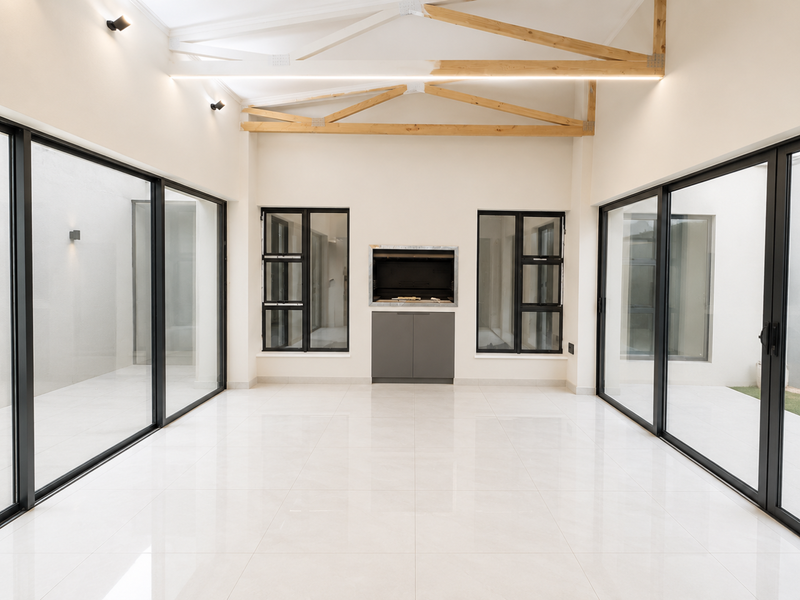 A bright, modern enclosed patio featuring exposed timber trusses, large format floor tiles, and a built-in braai, opening to the outdoors via sliding doors.