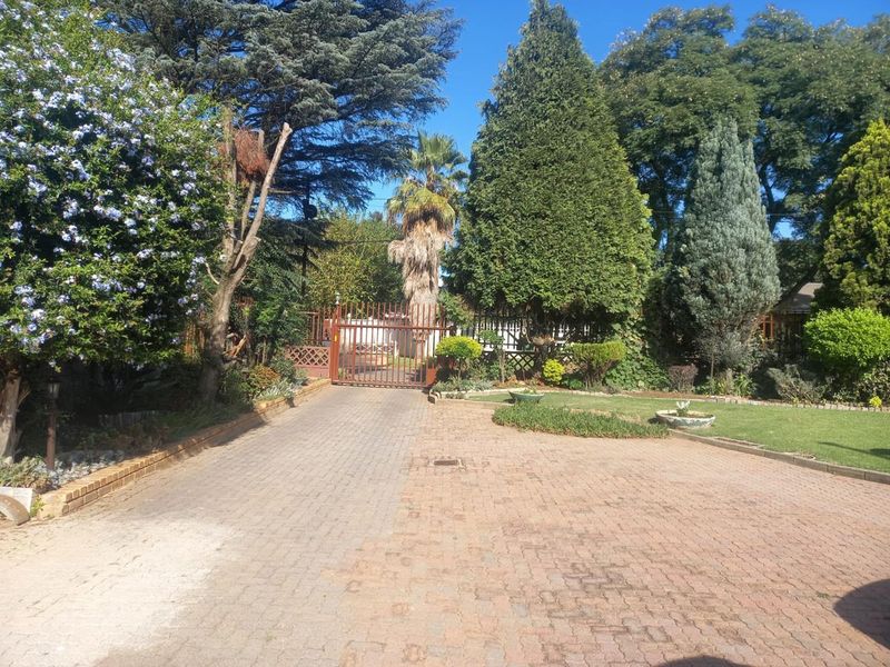 A well-maintained front entrance featuring a paved driveway leading to a secure metal gate, surrounded by established trees and a landscaped garden.