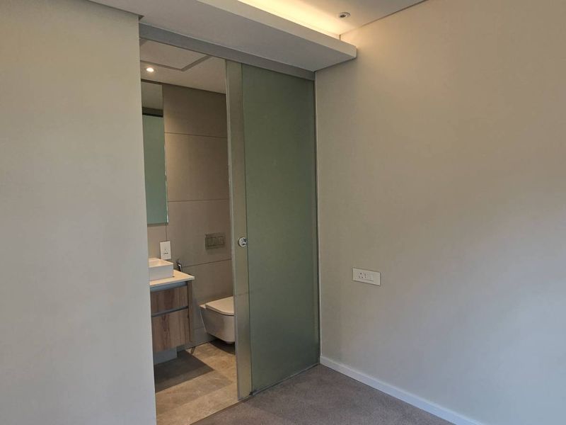 A clean, modern bedroom featuring a sliding glass door leading to an en-suite bathroom, neutral wall finishes, and recessed ceiling lighting.