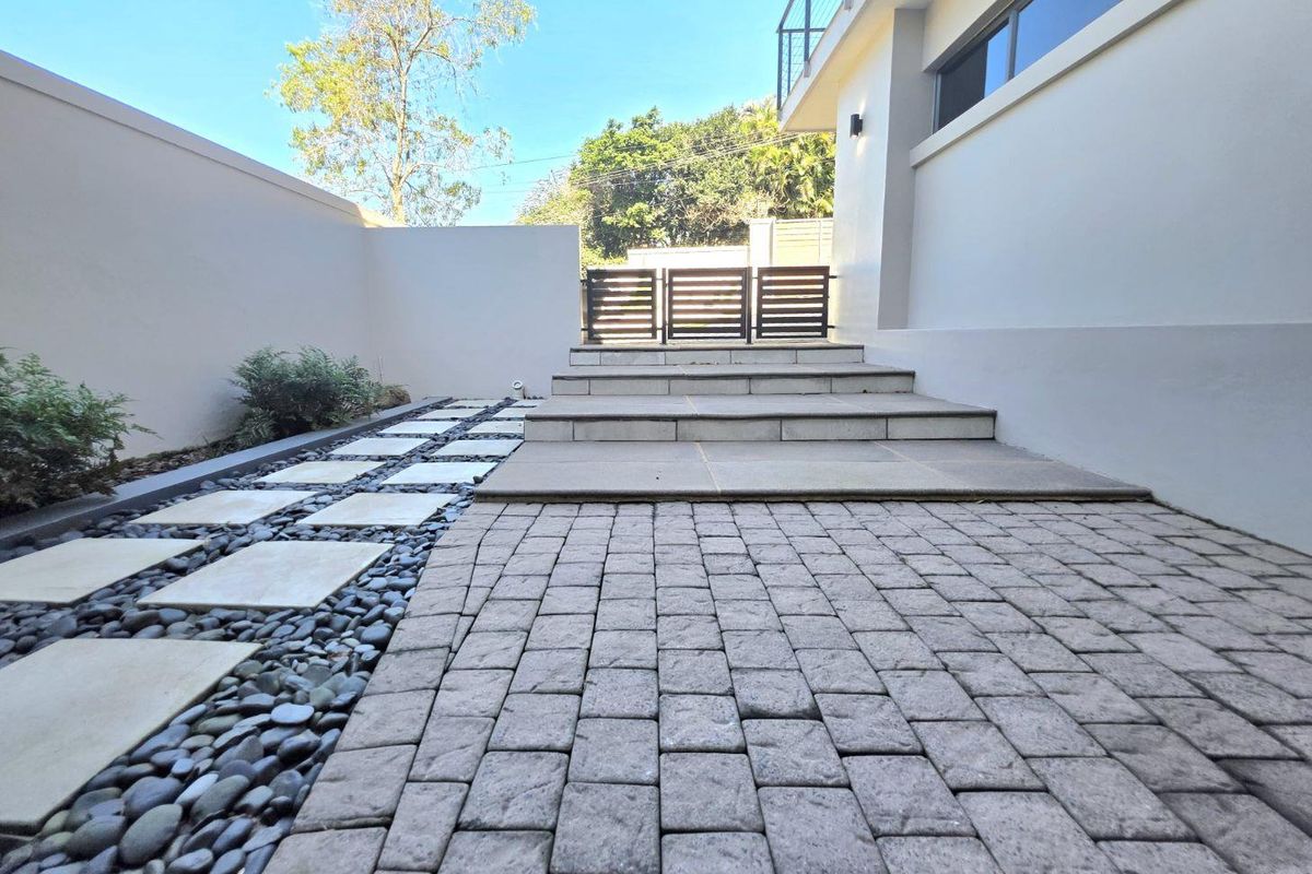 A well-maintained exterior rear courtyard featuring stone paving, a decorative pebble border, and concrete steps leading to a gated entrance.