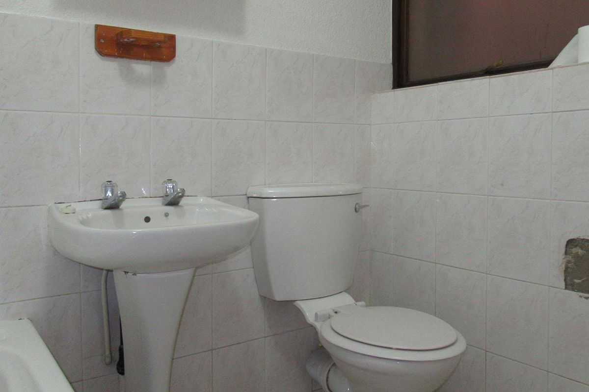 A functional bathroom featuring a pedestal basin and standard toilet, finished with white wall tiling and a small window.