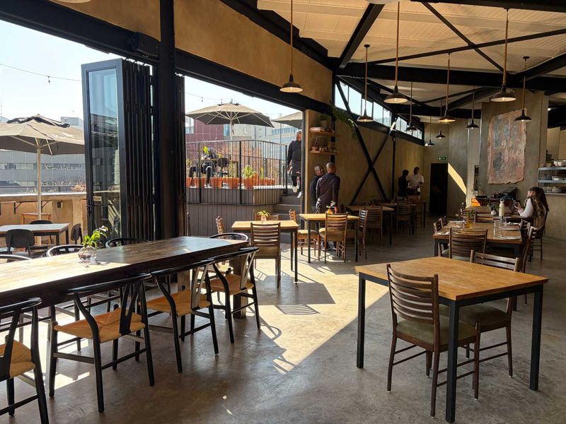 Spacious open-plan dining area featuring industrial-style exposed steel roof trusses, polished concrete floors, and large bi-fold doors opening onto an outdoor terrace.