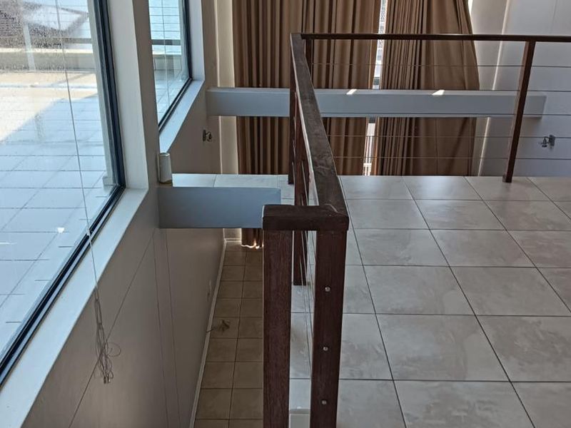 An open-plan landing area featuring tiled flooring, a wooden staircase railing, and large windows providing natural light and access to an outdoor balcony.