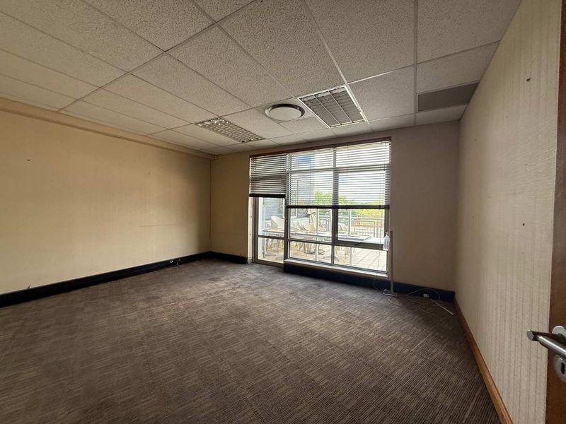 A vacant office space featuring a suspended ceiling, recessed lighting, carpeted flooring, and a large window providing natural light and access to a balcony.