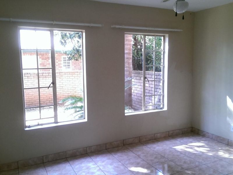 A bright, empty bedroom featuring tiled flooring, neutral walls, and two steel-framed windows overlooking the exterior area.