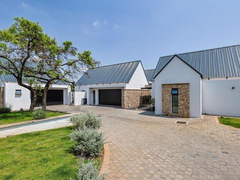 A well-maintained front exterior view featuring a paved driveway, stone-clad accents, metal roofing, and manicured garden areas leading to multiple garage bays.