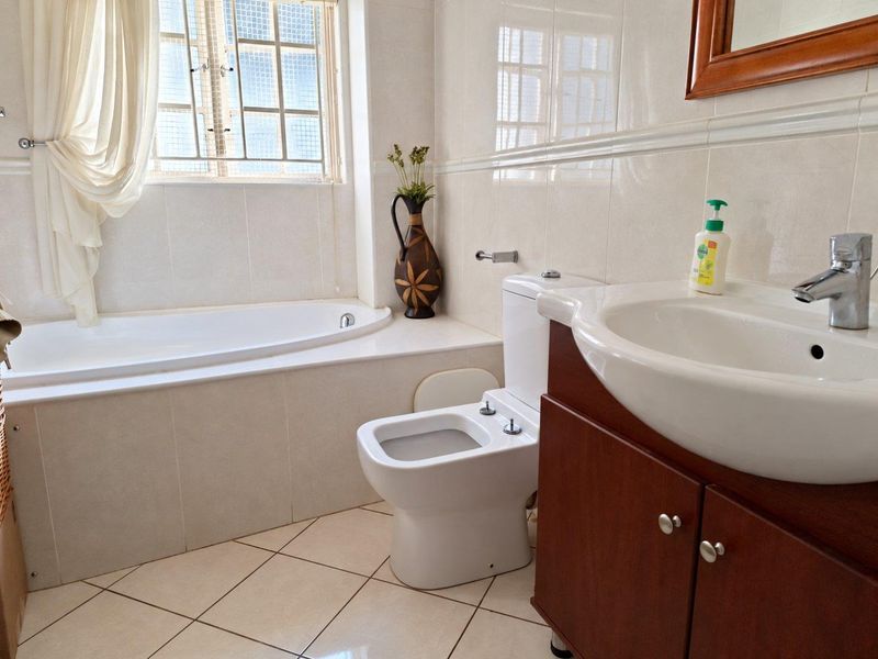 A functional bathroom featuring a built-in bathtub, a vanity cabinet with a basin, and a toilet, finished with neutral wall and floor tiles.