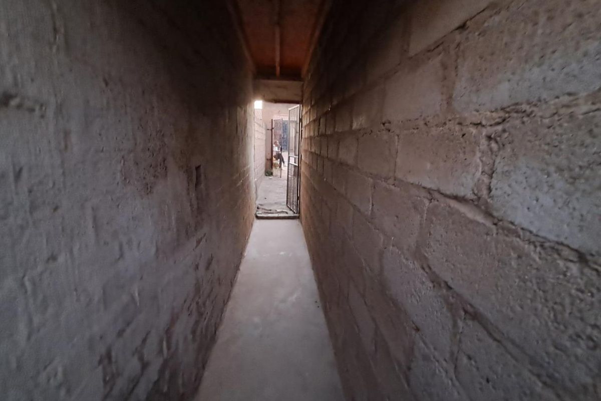 A narrow, unfinished exterior passageway featuring exposed concrete block walls and a concrete floor, leading to a metal security gate.
