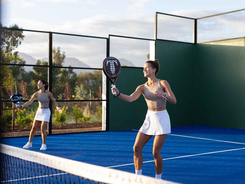 A well-maintained outdoor padel court featuring blue synthetic turf, perimeter fencing, and a central net, set within a landscaped residential environment.
