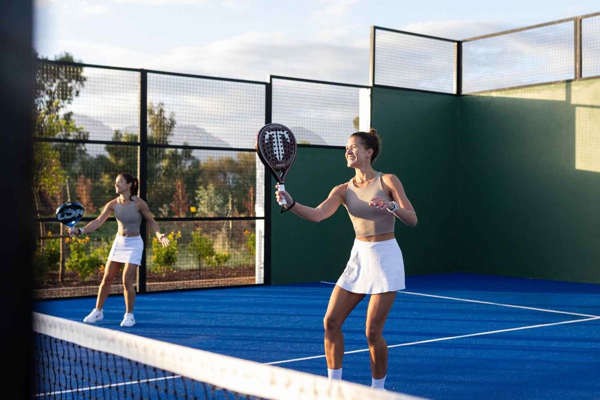 A well-maintained outdoor padel court featuring blue synthetic turf, perimeter fencing, and a central net, set within a landscaped residential environment.