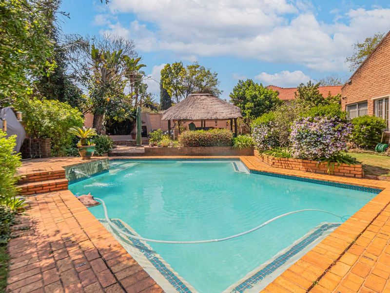 A well-maintained outdoor pool area featuring a swimming pool, brick paving, raised planter beds, and a traditional thatched lapa in the garden.