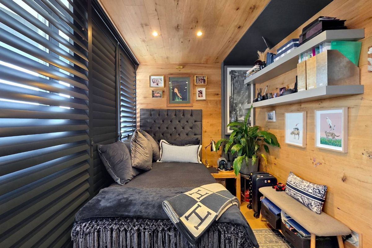 A cozy bedroom featuring warm wood-paneled walls and ceiling, integrated shelving, and dark adjustable window shutters.